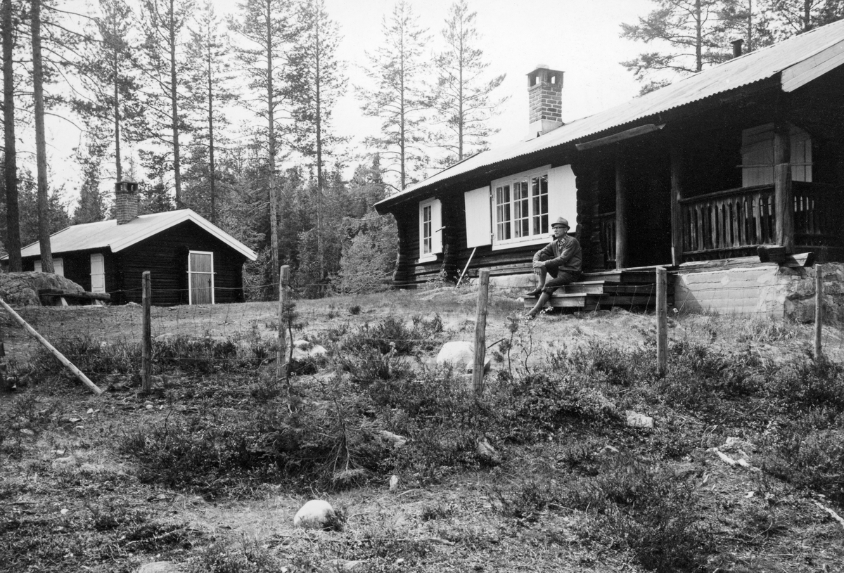 Kongsberg skogskoles praksissenter ved Sandoren i Hedenstad fondsskoger.  Tunanlegg med laftet våningshus til høyre og en mindre stuebygning til venstre.  En mann sitter på trappa foran den nærmeste bygningen.  Bygningene ligger på en bakkekam med furuvegetasjon. 