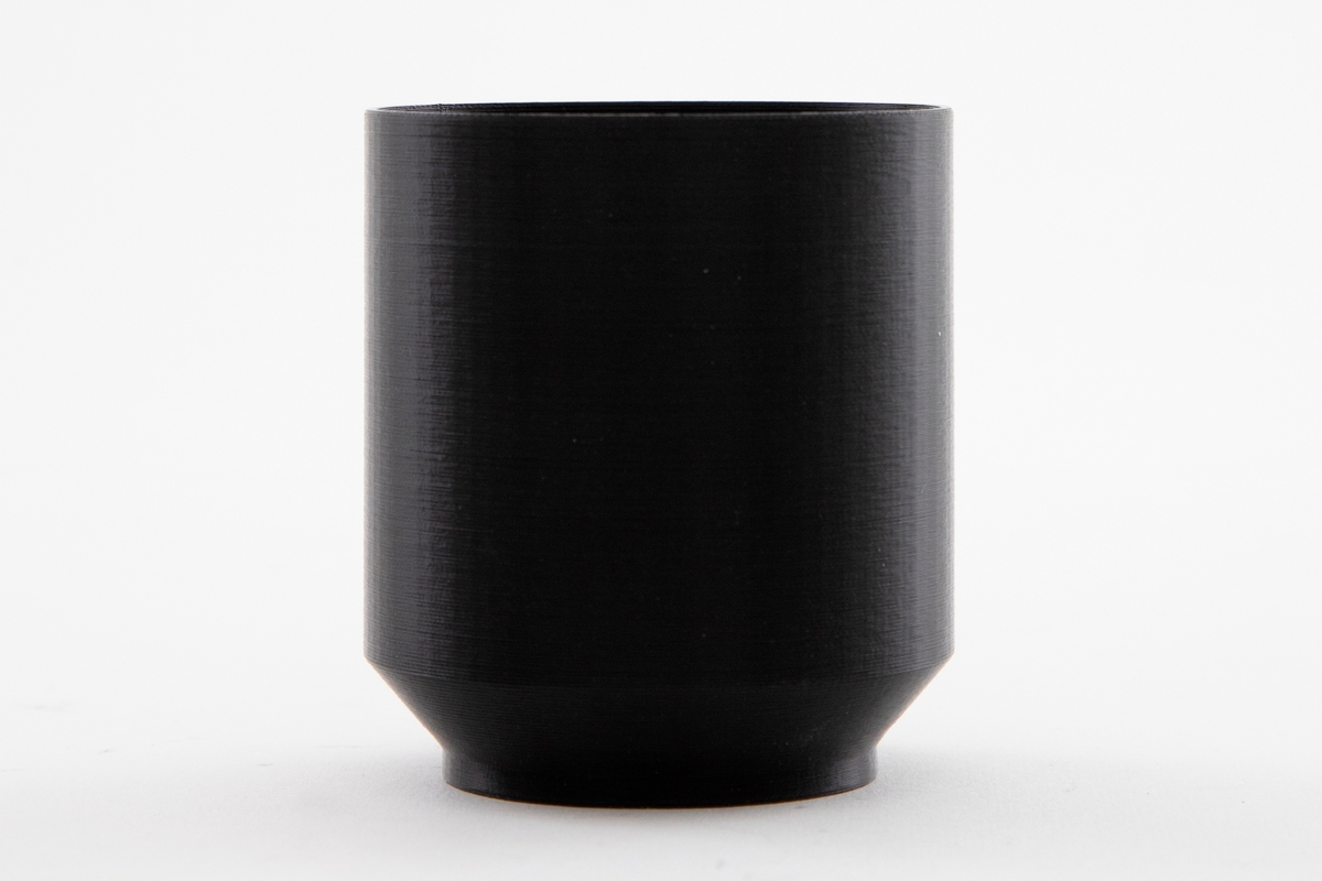 Liten og lav svart sylinderformet kopp. Sylinder brytes ved bunnen og skråner ned mot en liten rund fot.
