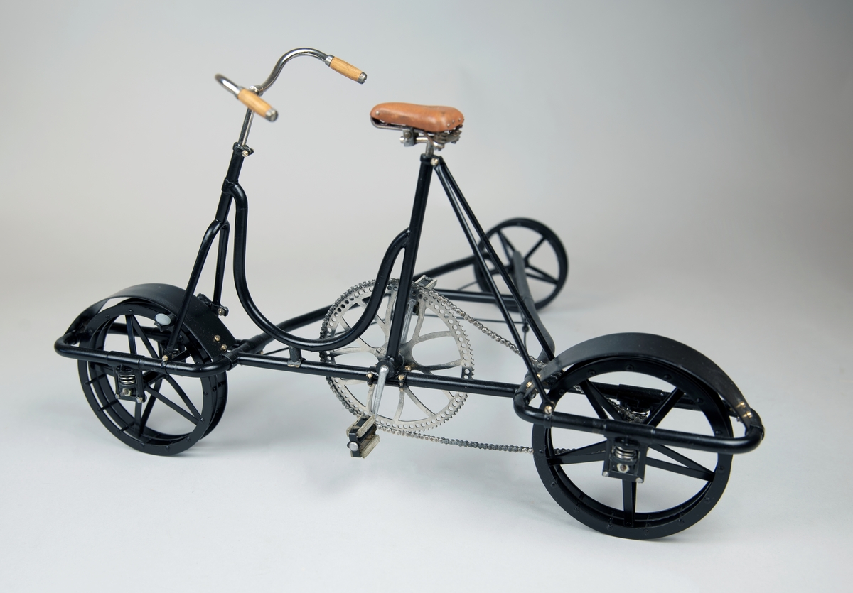 Modell av svart 3-hjulig cykeldressin, skala 1:5, av mässing och stål. Handtag i trä. Kedjan gjord av individuella länkar. Fjädring och broms fungerar. Ljusbrun sadel av formpressat läder.