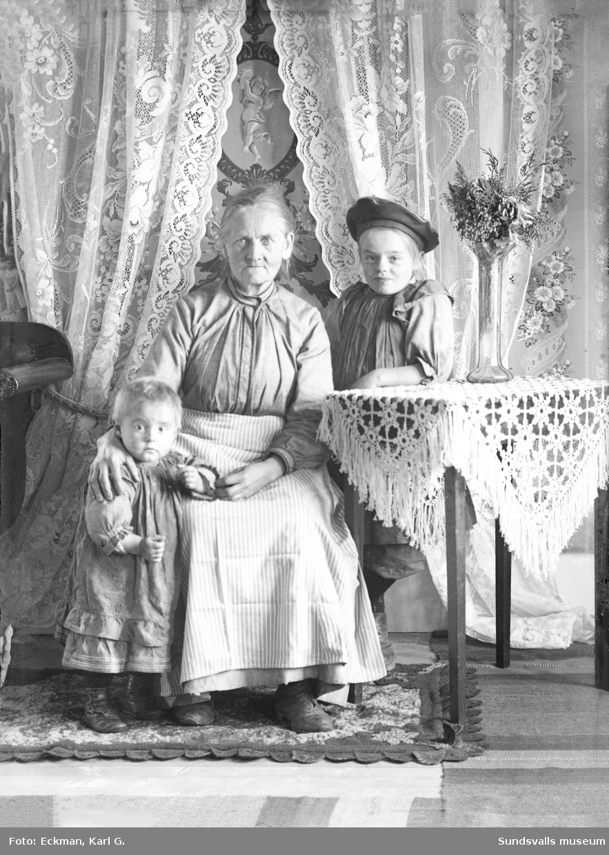 Porträttbild i hemmiljö med en äldre kvinna och två barn, Indal. Ur Karl Eckmans samling.