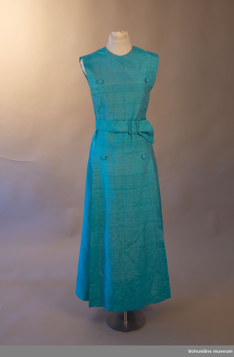 Turkos sidenklänning med ett hängande del framtill fästad i klänningens axel och halsdel. Tyget hålls in med skärpet som knytes i midjan. Skärpet bidrar till klänningens fall och form.