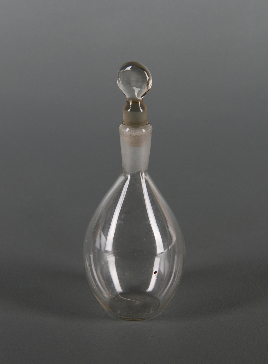 En flakong av glass, trolig borosilikatglass. Glasskolben har rundoval form (dråpeformet) med kort hals. Halsen er frostet. I halsen står det en glasspropp med rund topp.