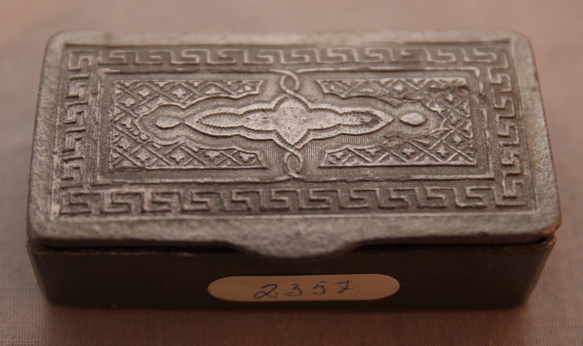 Fyrkantig dosa av bly. Locket har en inpräntad dekor föreställande en meanderbård samt ett orientaliskt mönster innuti denna.