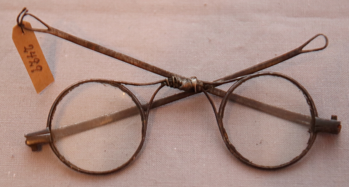 Glasögon med runda glas. Skalmarna är tillplattade och avslutas med en ögla som är avsedd för sammanbindande av ett snöre bakom huvudet. Näsbågen är skadad och lagad med en bit fastlödad ståltråd.