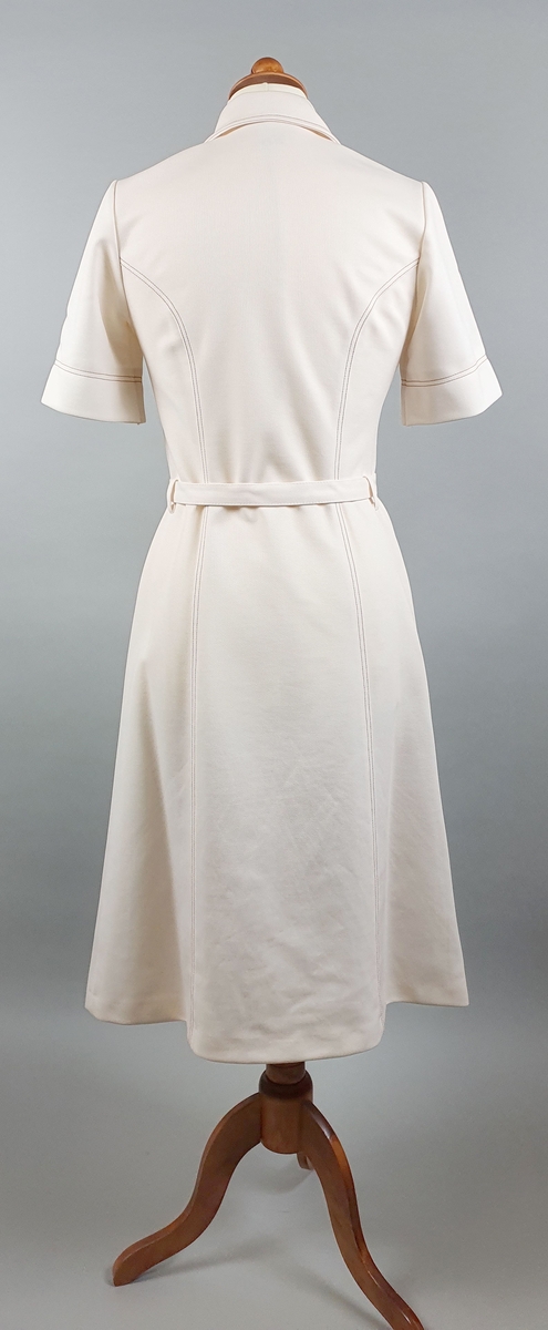 Hvit kjole med knapper foran, skjortekrage, påsydde lommer og belte. Brune dekorsømmer.