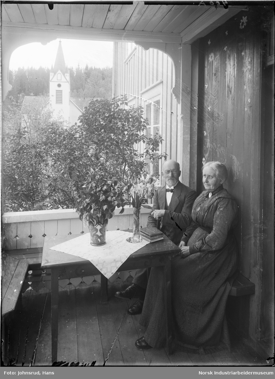 Par sittende på terassen til huset ved Nissedal prestegård. Nissedal kyrkje synlig i bakgrunnen.