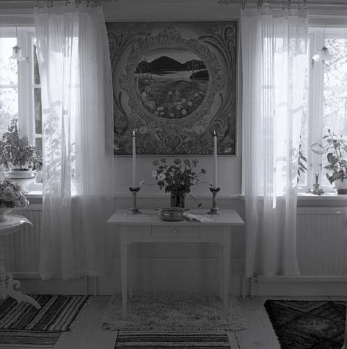 Lill-Annas dop sommaren 1973, här i ett rum på gården Sunnanåker. Där står ett vackert dukat bord med en blombukett, två ljusstakar coh en skål med dopvatten. Solen lyser in genom fönstrens skira vita gardiner.