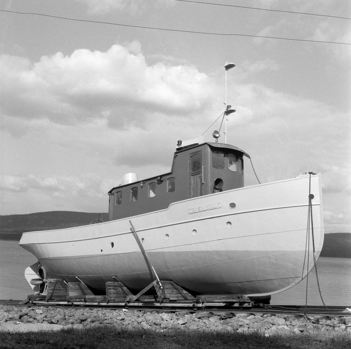 Slepebåten «Trysilknut», fotografert ved slippen på Sørlistøa ved Osensjøen i Åmot kommune i Hedmark i 1964. Fartøyet er fotografert skrått forfra, mot styrbord side. I 1964 var dette fartøyet 50 år gammelt. Skroget var slik det alltid hadde vært - 55 fot langt og 12 fot bredt, klinket sammen av det som i 1914 var «bedste sort skibsbygningsstaal», levert av Glommens mek. Verksted i Fredrikstad. Men Trysilknut var i utgangspunktet et dampdrevet fartøy. Dampmaskinen viste seg imidlertid i veikeste laget for de store tømmerbommene som skulle bukseres over den bortimot tre mil lange og 47 kvadratkilometer store Osensjøen. Etter 2. verdenskrig ble mange av slepebåtene som fellesfløtingsforeningene disponerte ombygd til dieseldrift. Ved å gjøre en slik investering så man for seg at man på litt lengre sikt skulle oppnå større trekkraft og samtidig redusere driftskostnadene. Dieselmotorene krevde nemlig langt mindre betjening enn dampmaskinene, og følgelig ville en dieseldrevet slepebåt kunne opereres med mindre mannskap enn et dampfartøy. I Trysilknuts tilfelle så man for seg at to fyrbøtere og en maskinist kunne utgå fra besetningen. Her lå det et betydelig innsparingspotensial i en periode med stigende lønnskostnader. I 1956 sendte direktøren i Glomma fellesfløtingsforening prisforespørsler til firmaer som man antok kunne påta seg ombyggingsarbeidet. Oppdraget gikk også denne gangen til Glommens mek. verksted. Demonteringa av dampmaskinen innebar også at det ble en del ledig plass om bord. Ominnredningsarbeidet ble overlatt til Ivar Hagens båtbyggeri på Hjelset utenfor Molde. Resultatet ble at Trysilknut - etter ombyggingsarbeidet vinteren 1957-58 framsto med et noe endret eksteriør og et fullstendig modernisert interiør.