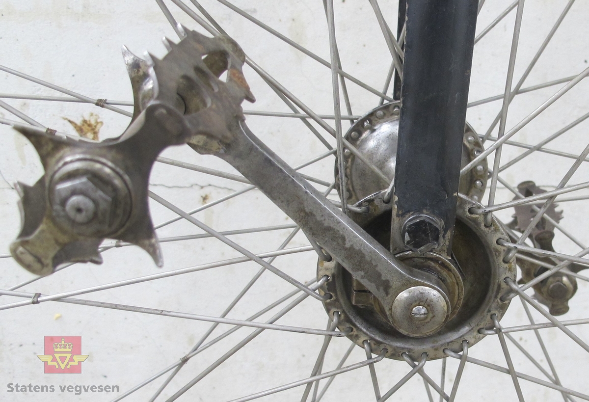 Sortlakkert velosiped med stort forhjul og lite bakhjul. Pedalene er montert direkte på fornavet og setet er plassert rett over. Hele sykkelen er av metall untatt håndtakene og dekk. Håndtakene er av tre og dekken av gummi.