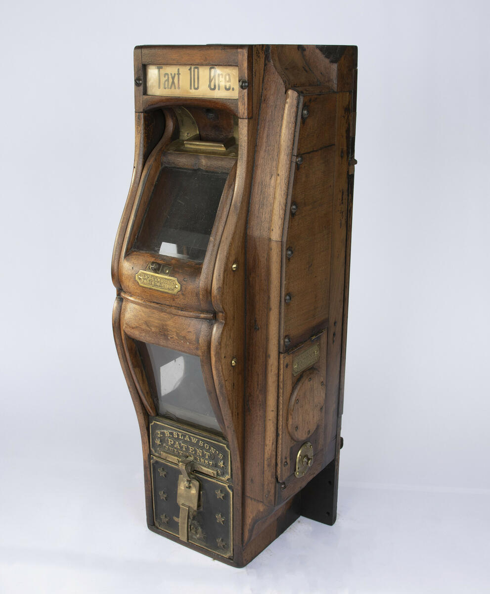 Betalingsautomat i mørkt treverk med vindu for kontroll av betalingen, safe i bunn og luke på siden for belysning av "kontrollrommet".