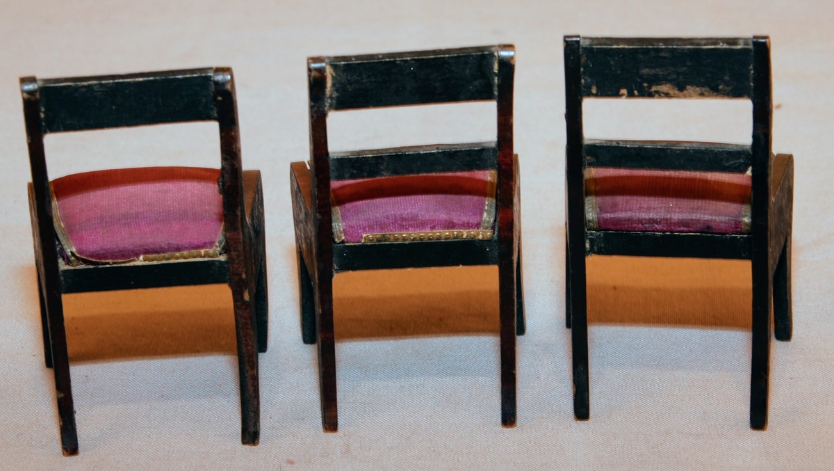 Tre stolar till dockskåp från mitten av 1800-talet i empirestil. I mörkbrunt trä med lila stoppade sitsar och guldiga pärlbårdar kring sitsarna. Med två tvärslåar i ryggen (en saknas på en av stolarna).