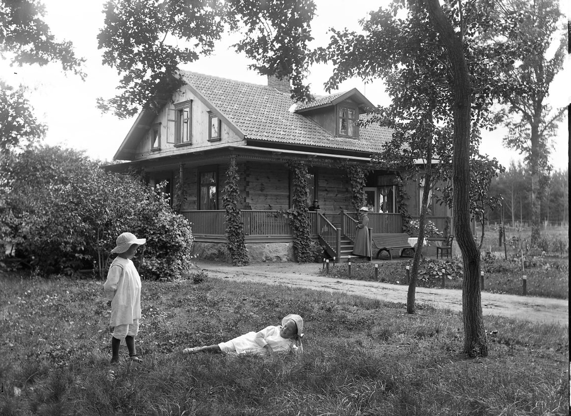 Två sommarklädda barn på en gräsmatta framför ett knuttimrad tvåvåningshus. På trappan till huset står en kvinna på och på verandan, som löper utmed framsida och gavel, sitter en kvinna.