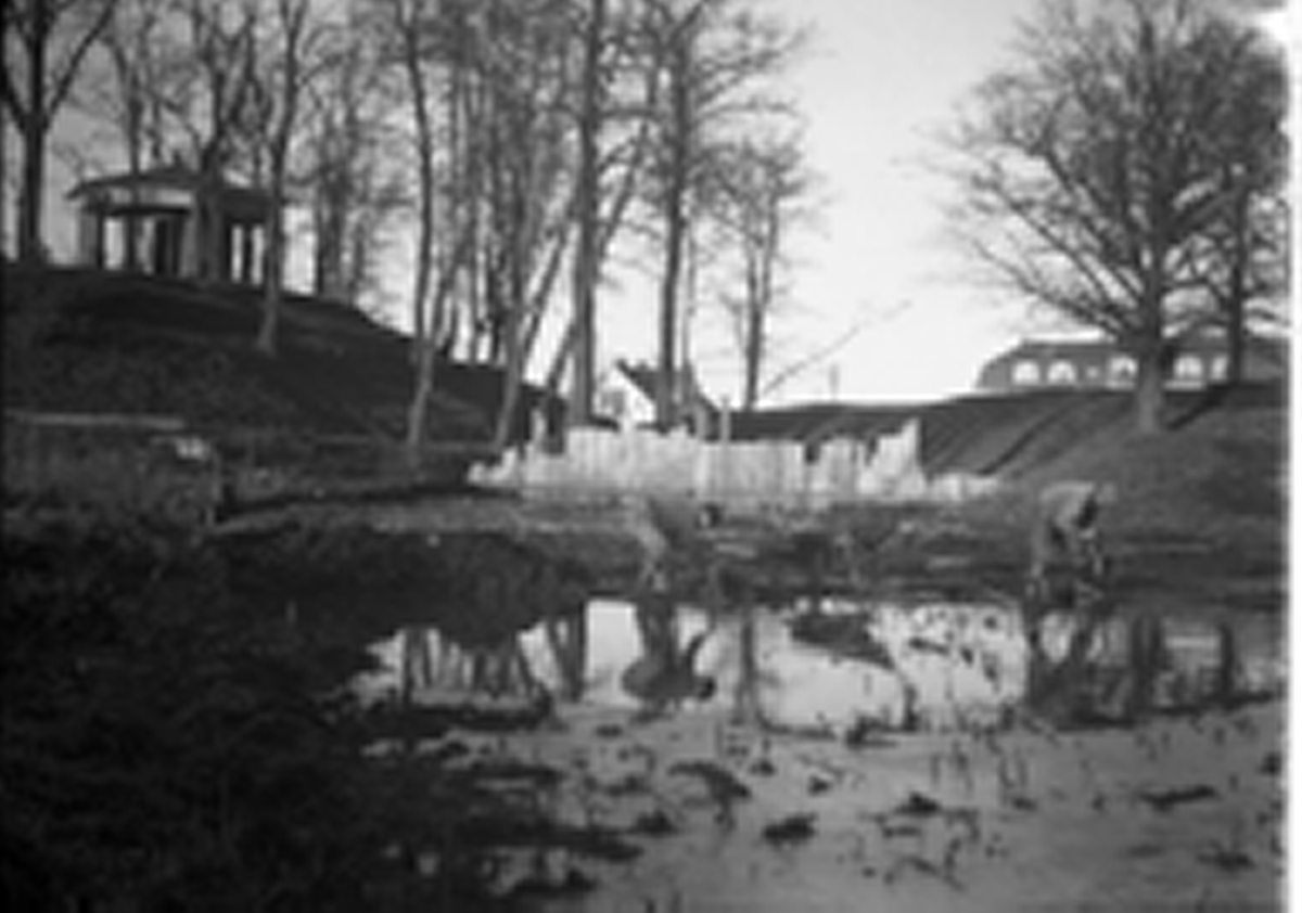 Två män arbetar med grävarbete i den inre vallgraven vid Rumlaborg i Huskvarna. Till vänster syns borgkullen med lusthuset på toppen och centralt en spåntvägg som har anlagts tvärs över kanalen för att hålla vatten borta. Fotot är taget i samband med arkeologiska undersökningar av den inre vallgraven, "kanalen", vintern 1934-1935.