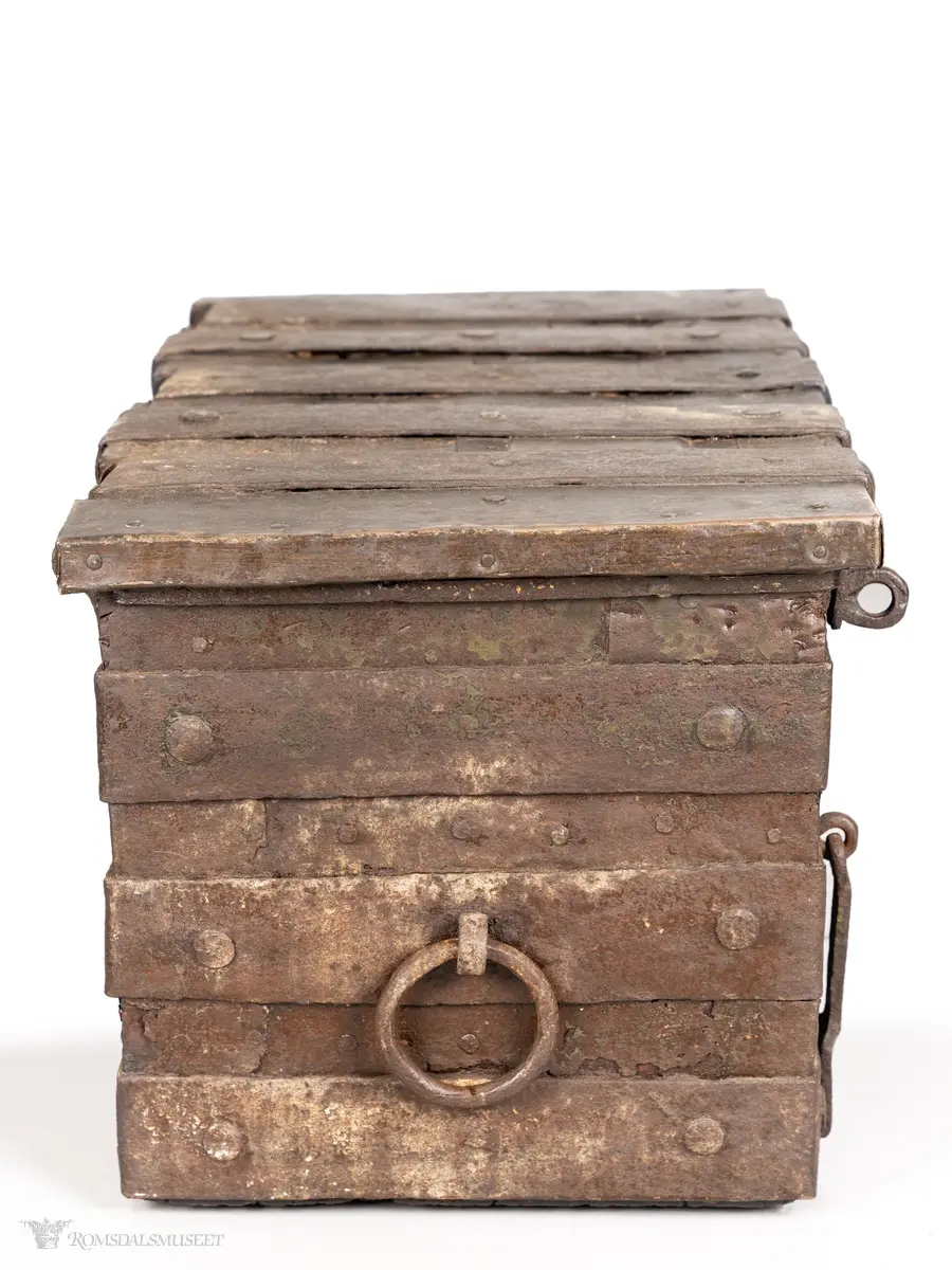 Solid kiste av tre, beslått med jernbeslag og jernplater. På høyre side er en jern- ring som benyttes som en slags nøkkel i tillegg til en annen nøkkel.