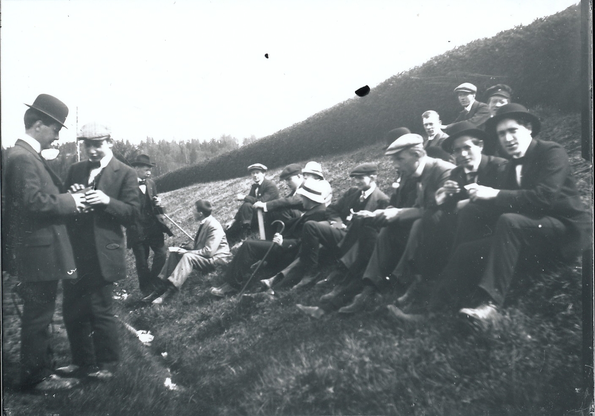 En gruppe yngre menn sitter og står i graskledd skråning. Hekk øverst i skråningen.