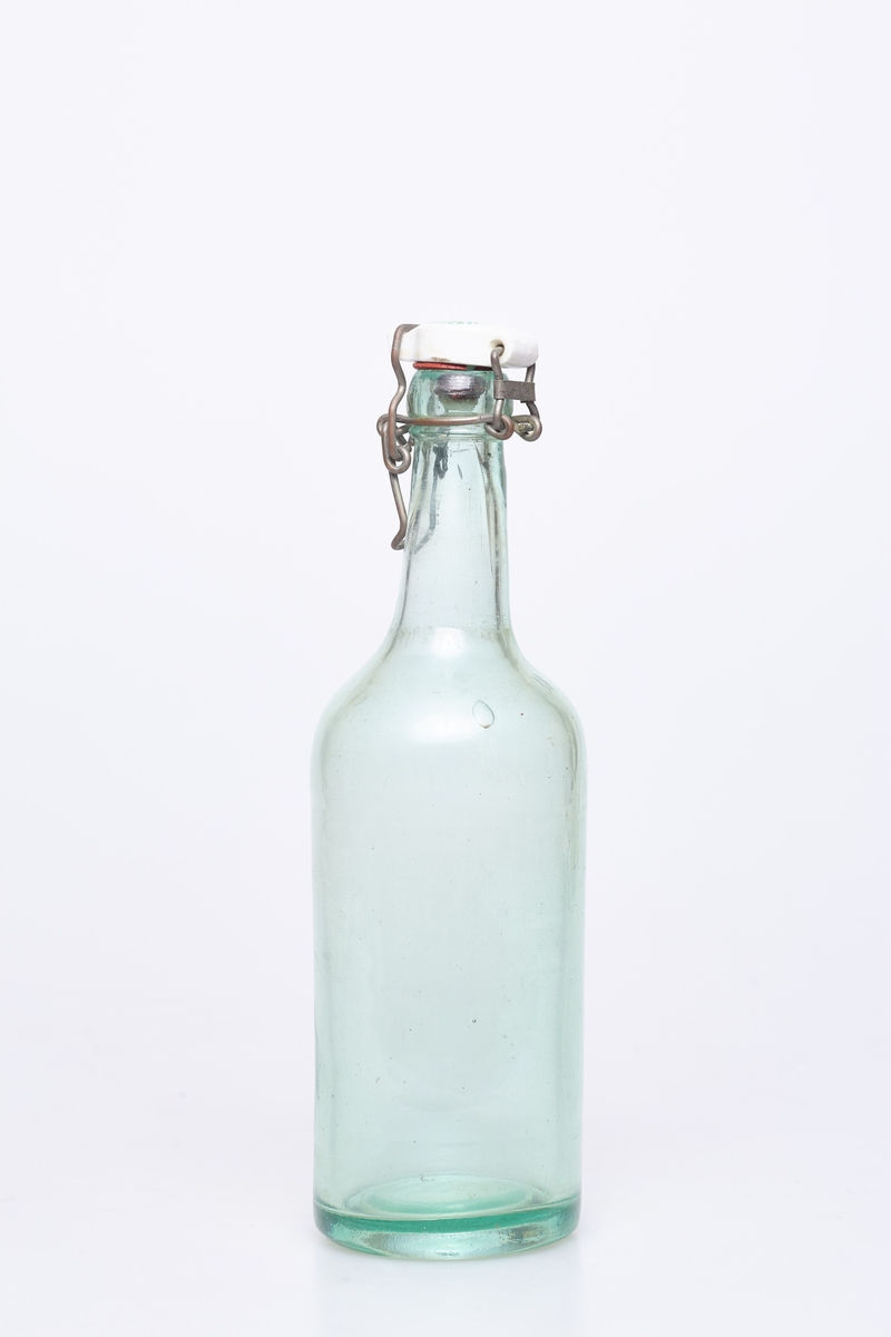 Glassflaske av klart glass med noe grønnskjær. Har hvit plastkork med metallfeste.
