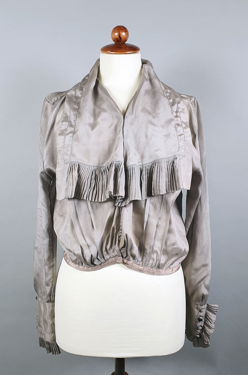 Gråbrun silkebluse med jakkeslag med rysjer langs kanten og over brystet. Blusen lukkes med trykknapper. Lange ermer med mansjetter med knapper og rysjekant.