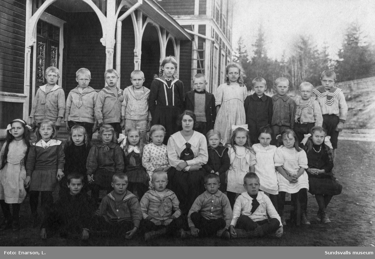 Skolfoto med en kvinnlig lärare och 27 barn utomhus framför en lantligt belägen skola. Ur en samling från Fanbyn, Stöde.