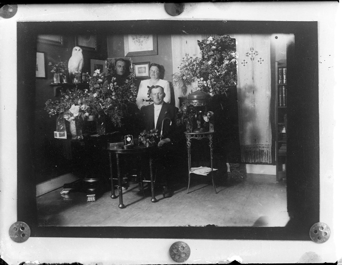 Interiörbild. En man sitter vid ett bord med blommor. Troligen mannens födelsedag. Bakom honom står hans hustru och intill henne står ett porträtt/fotografi av en ung man - sonen Eric Alming 
Till höger en uppstoppad fjälluggla, möjligen.