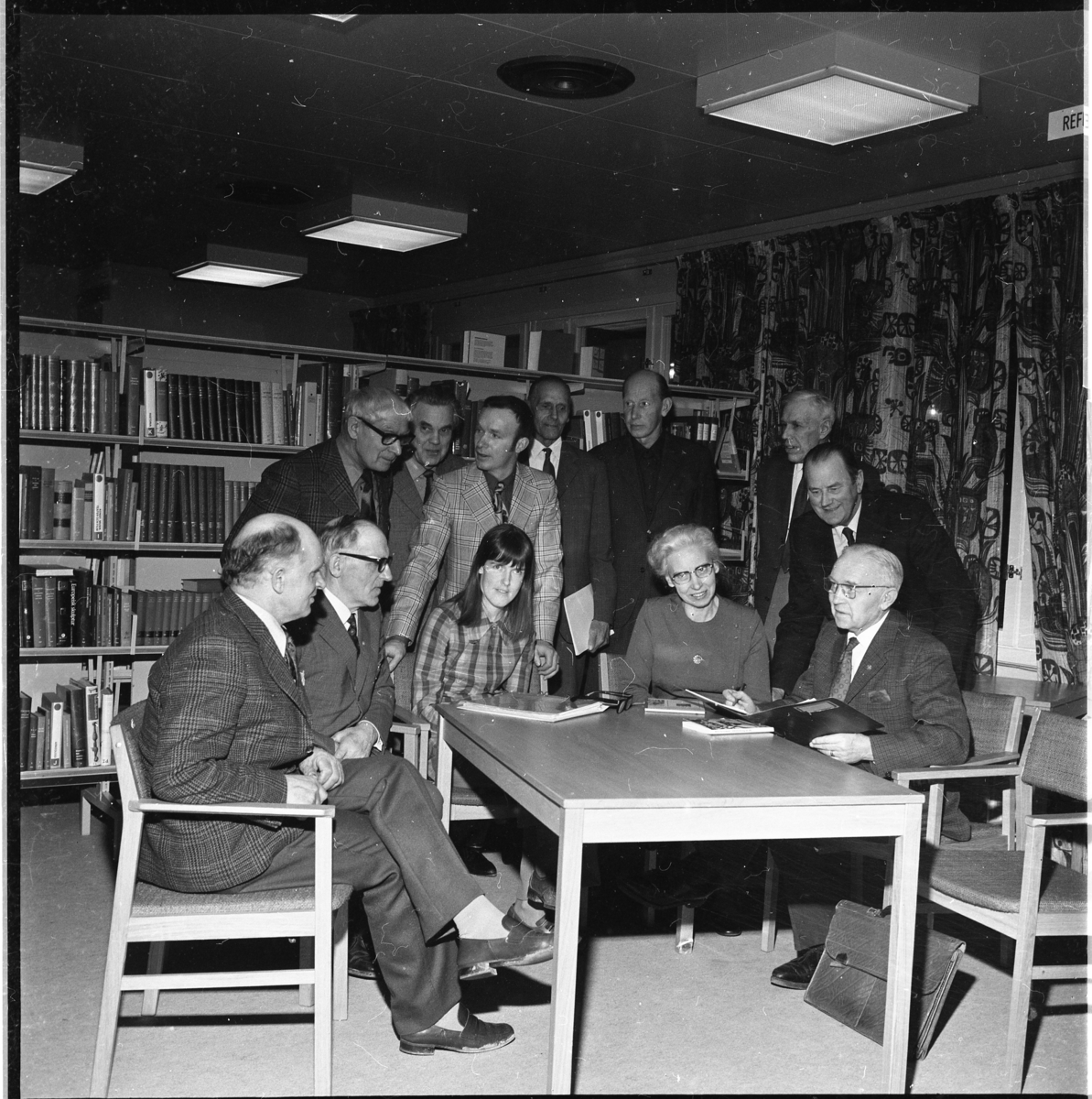 Två kvinnor och tio män stående och sittande runt ett bord. Fokus är riktat mot en man sittande till höger: Sven Simonsson. Stående som två från vänster är Sten Grennfelt. Kvinnan till höger är Astrid Wiråker och bakom henne står hennes man sågverksägaren Bengt Wiråker. 
Det är släktforskning, möjligen en kurs, under ledning av Sven Simonsson. De befinner sig möjligen i dåvarande biblioteket i Grenna Medborgargård.