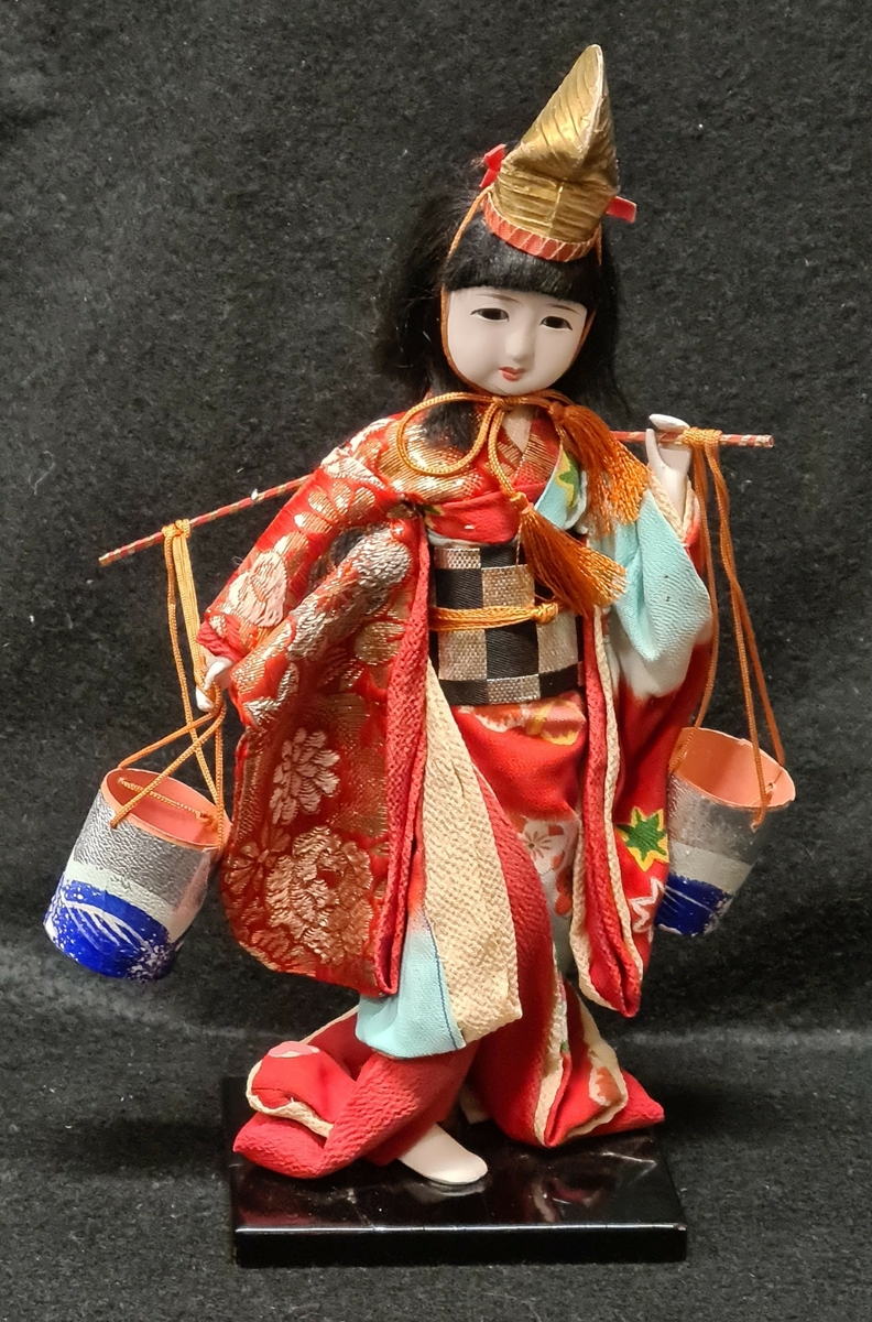 Docka i traditionell dräkt som tillhör Vera Hanssons docksamling.

Köpt 1959. Yoriko. Japan.
Japanska i mångfärgade kläder huvusakligen rött.
Hon bär ett ok med två ämbar.