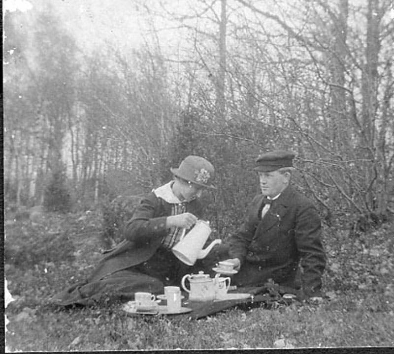 John Algot Carlsson på utflykt i en skogsdunge med kvinnligt sällskap i vårhatt. De har dukat upp med porslinsservisen och hon häller upp åt honom ur kaffekannan. Omkring 1915.