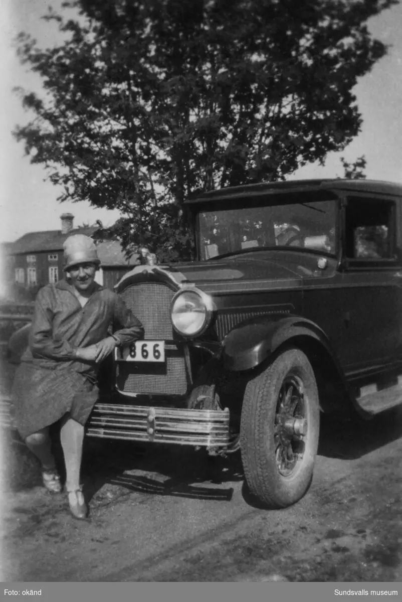 På kofångaren till en bil av äldre modell med registreringsnummer Y866, poserar en kvinna med hatt. Lantligt beläget bostadshus i bakgrunden. Ur en samling som tillhört Rodin/Öhlén, Kovland.