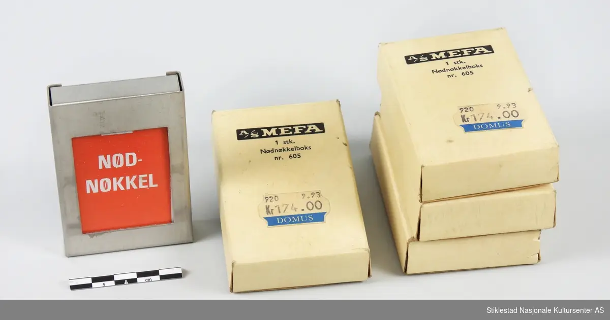 Eske/emballasje i papp merket A/S MEFA 1 stk. Nødnøkkelboks nr. 605.  Selve boksen er i stål med rød papirskilt i front merket NØD-NØKKEL. Priset til 174 kr. (DOMUS). 4 esker med nøkkelboks.