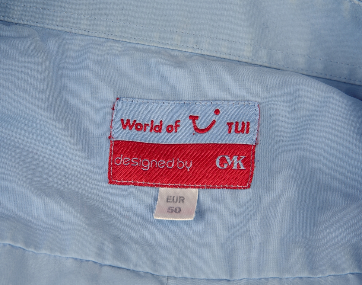 Ljusblå kortärmad skjorta i storlek 50. Skjortan är gjord i 55% bomull och 45% polyester. Knäpps med sju ljusblå knappar av plast på framsidan, två extra knappar finns längst ner på insidan av skjortan. En bröstficka på vänster sida. På vänster skjortärm finns TUI:s logotyp broderad i rött. En etikett i nacken där det står "World of TUI designed by GMK EUR 50". En tyglapp på insidan av skjortan med information om material samt tvättråd.