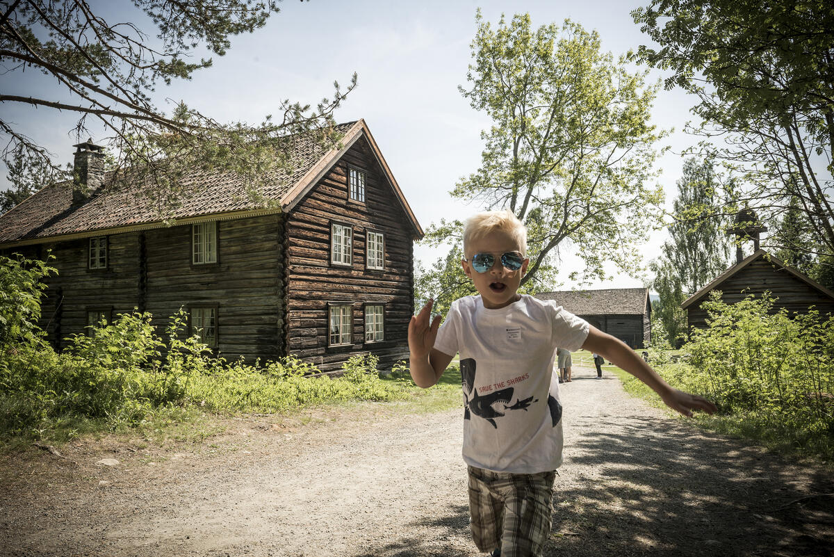 En gutt med solbriller løper mot kameraet. I bakgrunnen en gammel tømmerbygning. Sol og grønne blader på trærne.