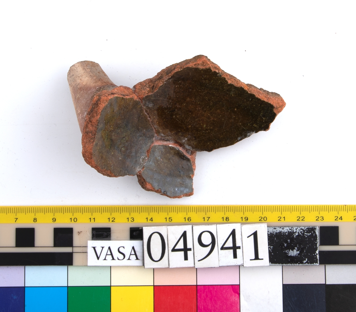 En keramikskärva med bit av ben (avslaget) H 50 mm. Fragment med bit av botten (hopsatt av 3 bitar). Insida av botten blyglaserad grönbrun. Utsida oglaserad med fläckar av glasyr.