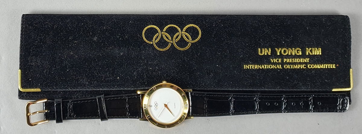 Gull- og hvitfarget armbåndsur med sort reim. På urskiven er det motiv av de olympiske ringene. Uret ligger i en svart, smal konvolutt, med olympiske ringer og tekst i gullfarge.