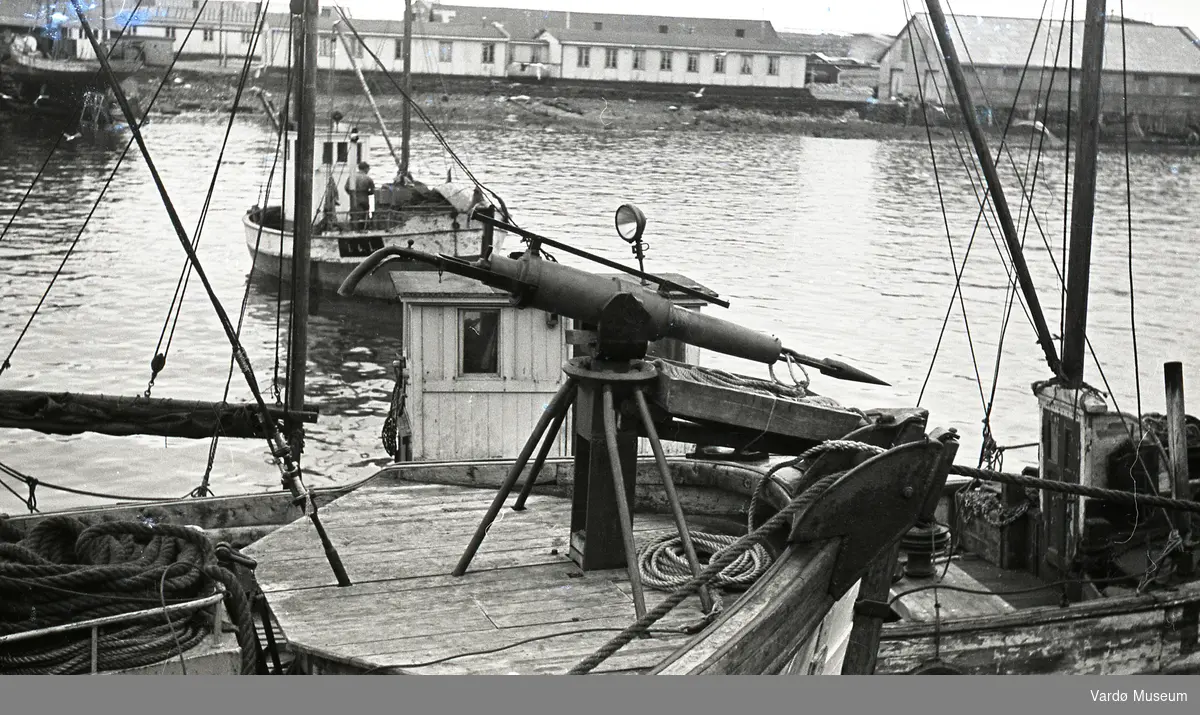 Harpun på en fangstbåt i Vardø, Vardø slipp i bakgrunnen samt internatboliger for Statens fagskole for fiskeindustri. Bygget øverst i høyre hjørne var en Guanofabrikk.