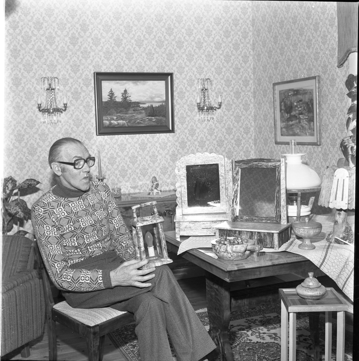 En man sitter i sitt hem omgiven av föremål, bland annat pigtittare. skålar och lampor. Förmodligen tillverkade av honom själv.