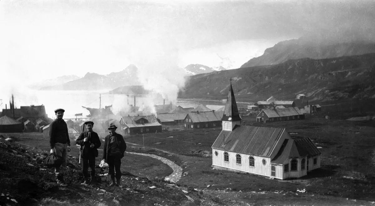 Sort-hvit-bildet viser tre menn oppstilt og et nakent landskap med hvit kirke i bakgrunnen. Lenger ned mot sjøen er noen arbeidsbygninger, båter og røyk eller damp.