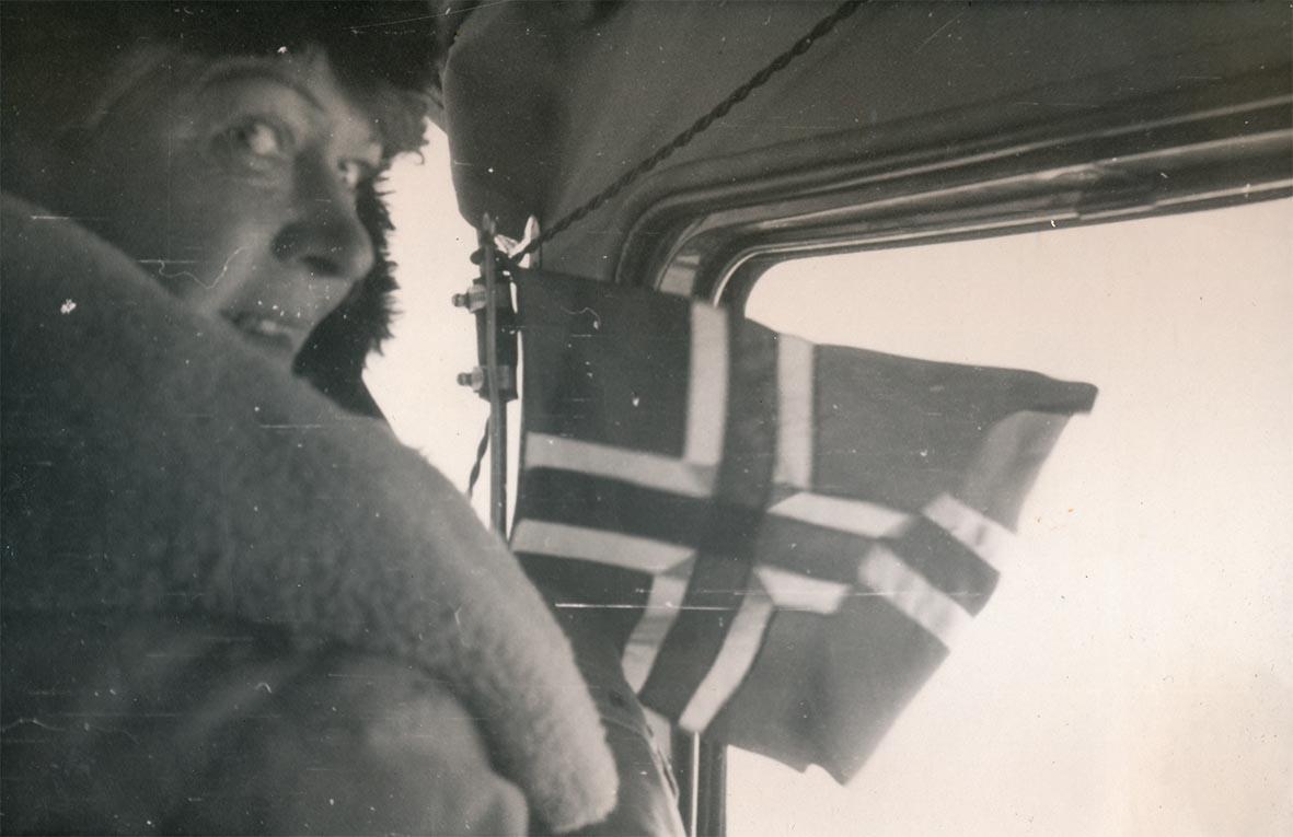 Sort-hvit-bilde viser en kvinne som ser mot oss over skulderen sin. Hun er kledd i pels og hatt og har et flagg foran seg. (Foto/Photo)