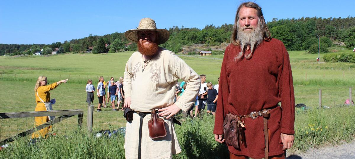 Bildet viser to vikingkledde menn med skjegg. I bakgrunnen er det grønne sletter, blå himmel og noen mennesker i aktivitet