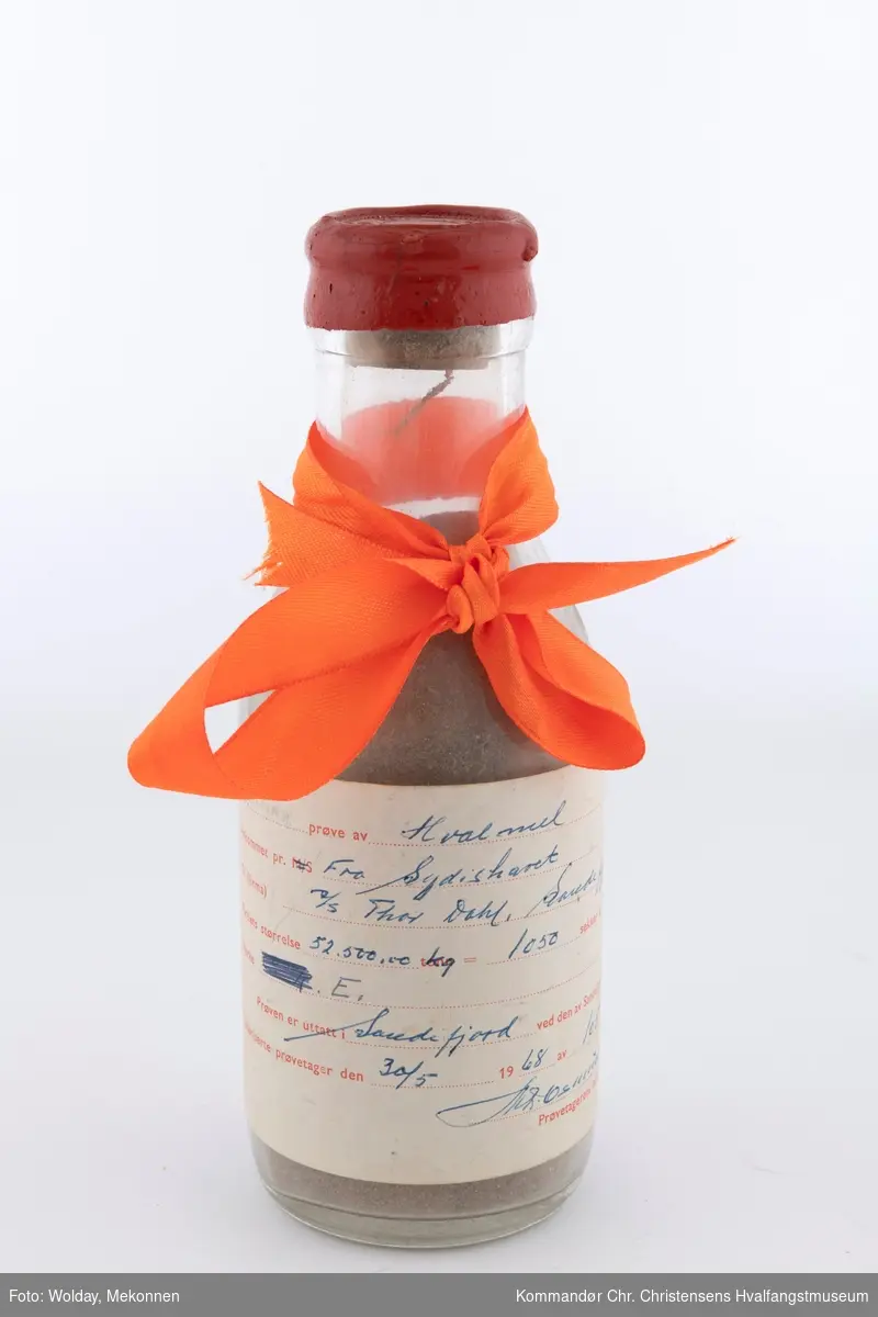 Glassflaske med etikett og vokset kort. Oransje gavebånd rundt flaskehalsen.
Flaske med prøve av hvalmel