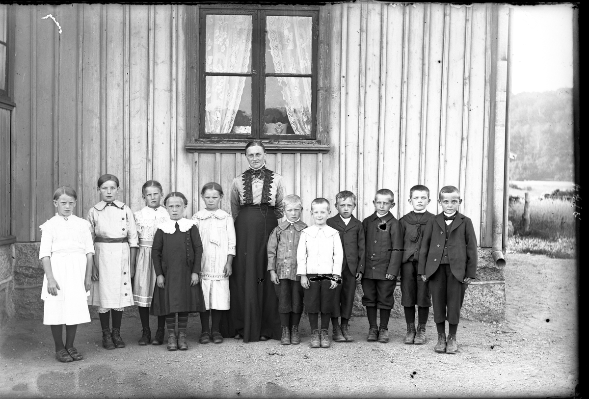 Småskollärarinnan Bertha Engström (fotografens mor) med en skolklass om fem flickor och sex pojkar. På den tiden ingick barn av olika åldrar i samma klass. Flickan i mörk klänning kallades Vävars-Ester (f. 1905); till höger om Bertha i randig kolt står Gade-John (f. 1906); som nr 4 står Ivar i Råryd (f. 1905); nr 5 är Gunnar i Kullen (f. 1906) och längst till höger står Sves-Gunnar (f. 1904).
