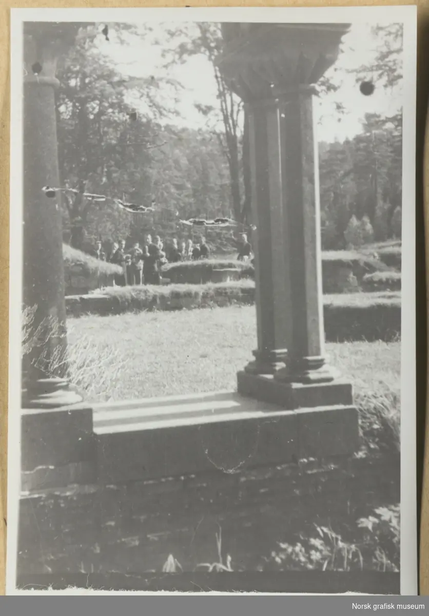 Utendørsmotiv. Steinsøyler i forgrunnen, i bakgrunnen skimtes en gruppe mennesker i et landskap. Fra Lysekloster. Fotografert i forbindelse med Vestlandsk Trykkerstevne i Bergen, 1946.