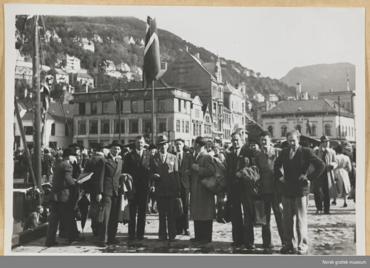 Gruppebilde i bylandskap. Menn i dress i forgrunnen, bybebyggelse i bakgrunnen, blant annet Norges Bank og Holberg-statuen. Fotografert i forbindelse med Vestlandsk Trykkerstevne i Bergen, 1946.