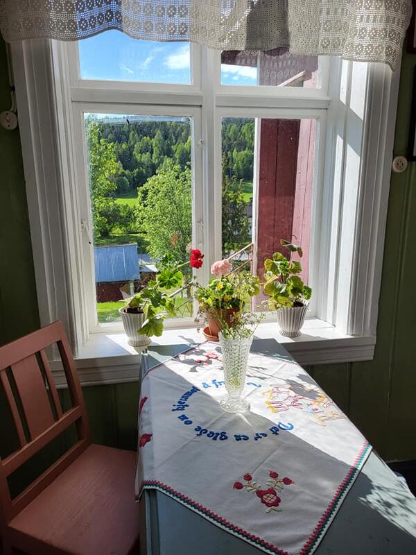 Kjøkkenet i Vestun på Eidskog bygdetun Almenninga. Grønn vegg, lyserød stol og lyseblått bord. Bord med brodert duk. Glassvase med villblomster på bordet. Hvit vinduskarm med tre blomsterpotter. I pottene er det røde og rosa pelargonier.