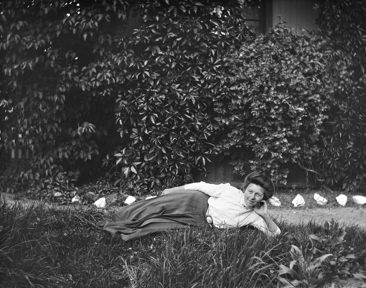 Svart-hvit foto av kvinne som ligger på siden i gresset. Hun hviler hodet i hånden, og har på seg hvit skjorte og skjørt.