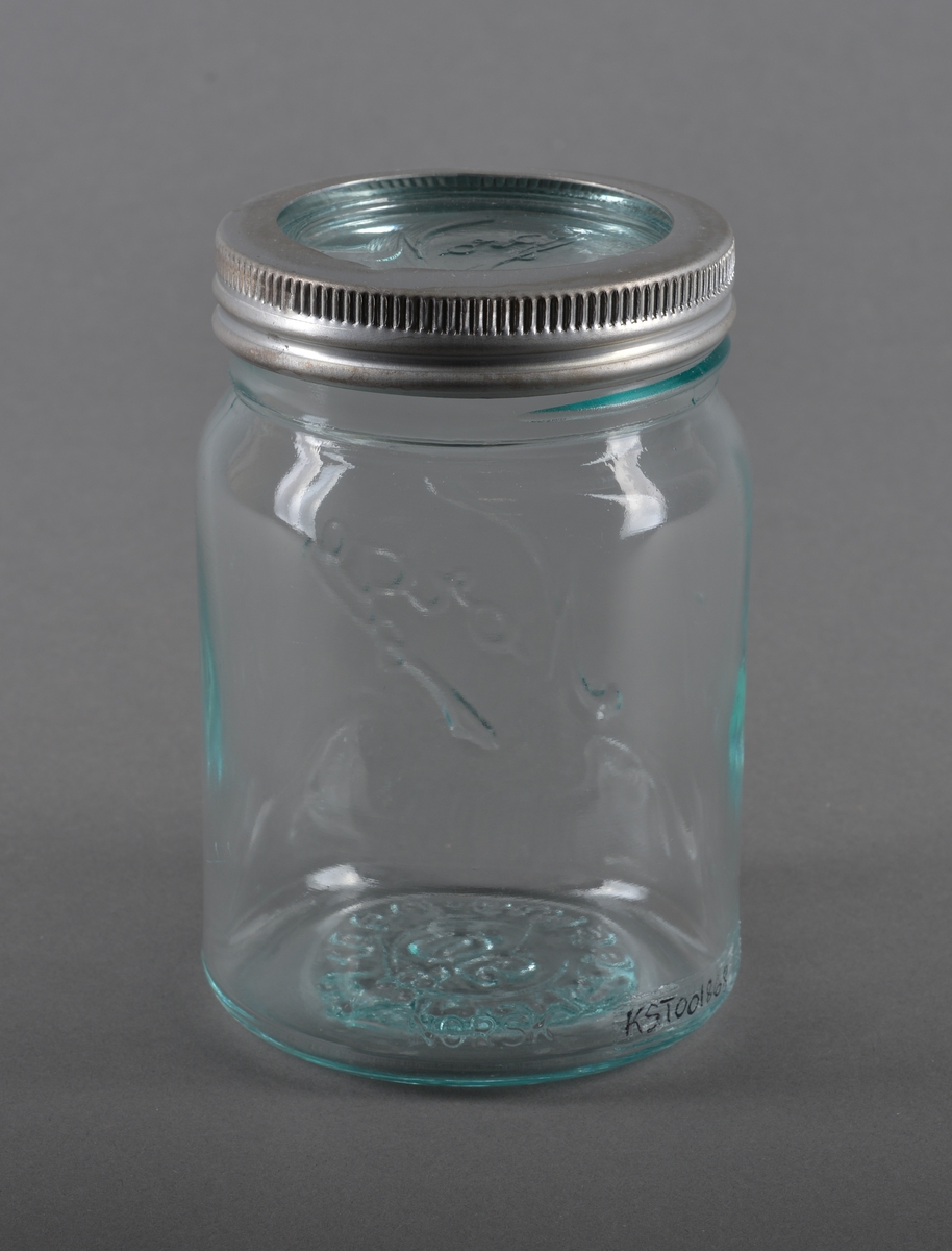 Sylindrisk glassbeholder med glasslokk, som har gjenget metallring. Glasset er gjennomsiktig.