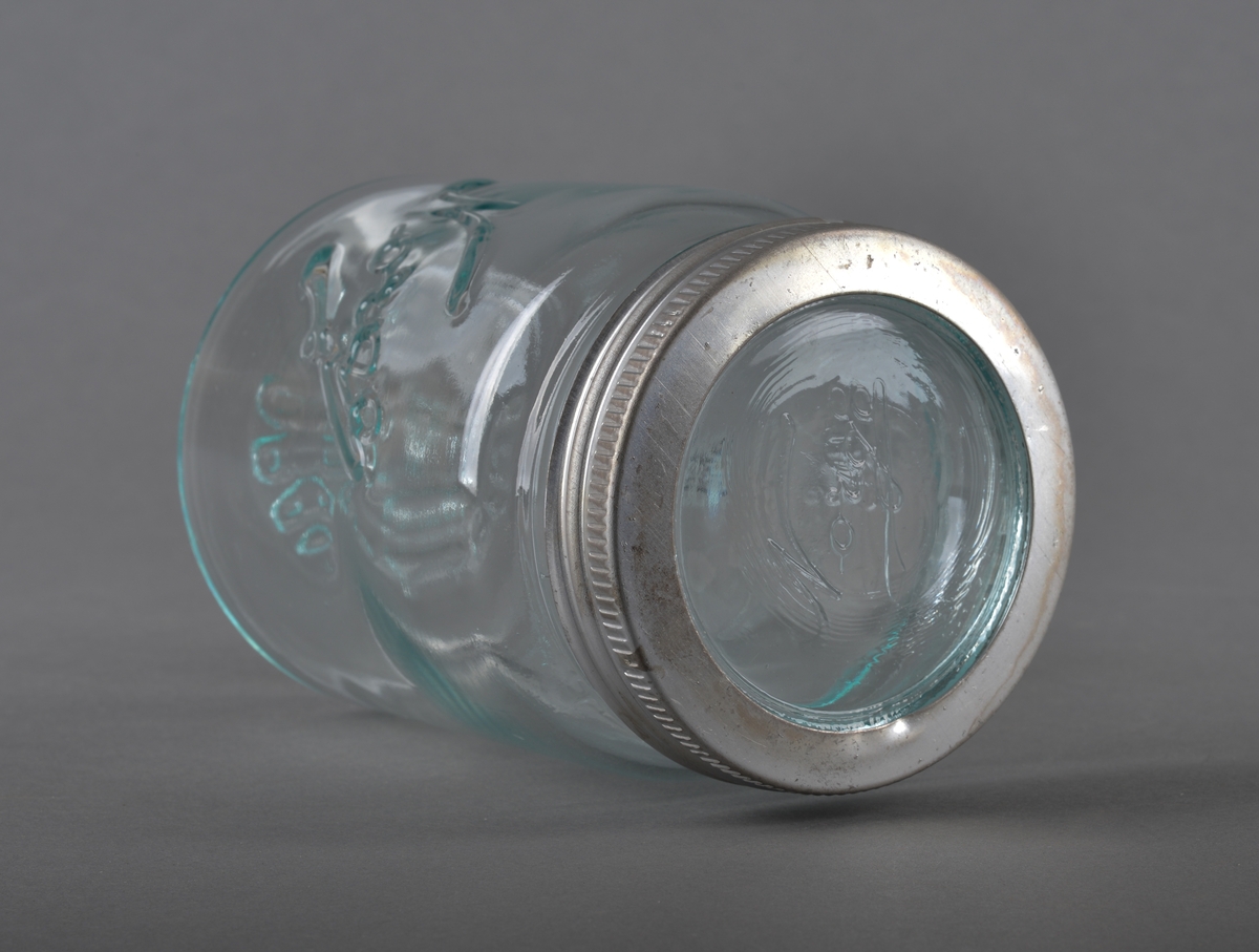 Sylindrisk glassbeholder med glasslokk, som har gjenget metallring. Glasset er gjennomsiktig.