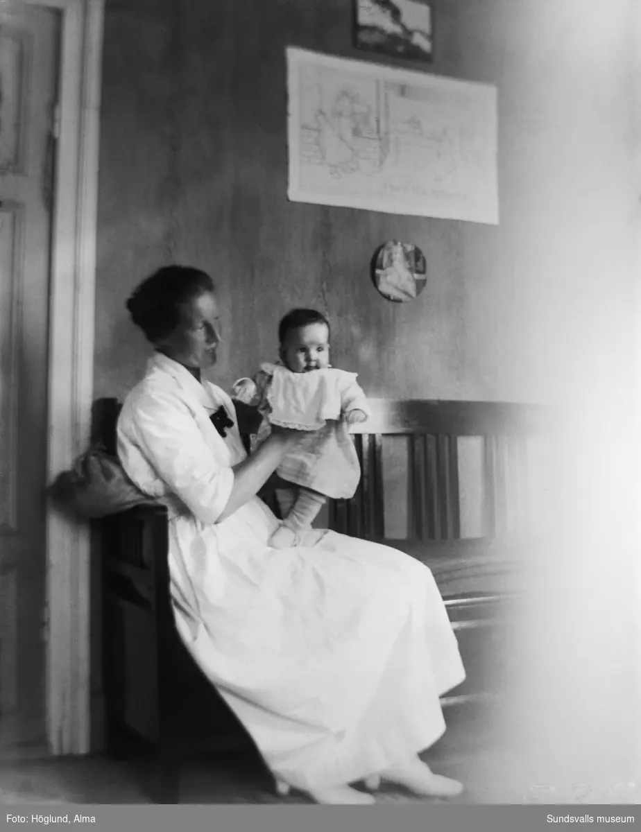 Porträtt i hemmiljö, en kvinna sitter i en kökssoffa med en baby i famnen.