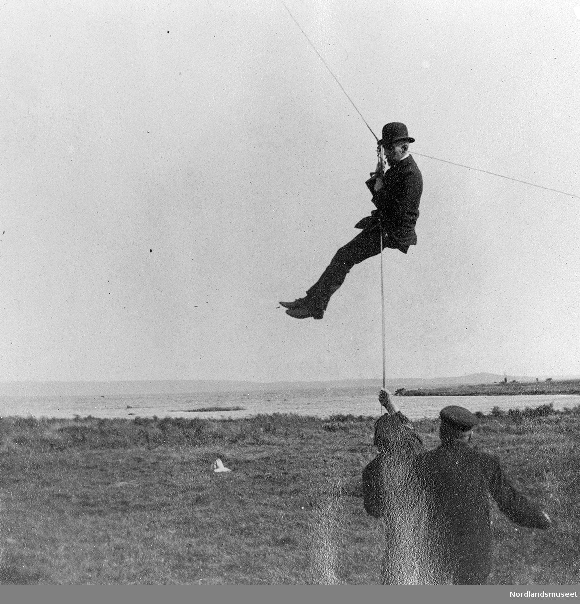 Drake, eksperiment/forsøk med draker. Roald Amundsen er en av deltakerne. To personer står på bakken og holder i et tau. Dette tauet er festet til lina der drakene er tilsluttet. Roald Amundsen holder seg fast der tauet og drakelina er festet sammen, slik at han befinner seg ca 2 - 3 meter oppe i luften.