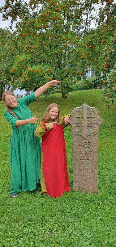 To unge jenter i middelalderkjoler står og peker på en pilegrimsmilestein med påskriften "488 km til Nidaros" (Foto/Photo)