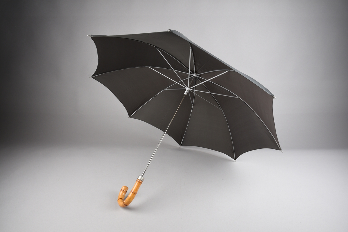 Paraply med 8 spilar og stong i metall. Handtak i bambus. Paraplyen har varetrekk som vert tredd innpå paraplyen, enden på stongen stikk ut enden. Knepping for lukking av trekket.