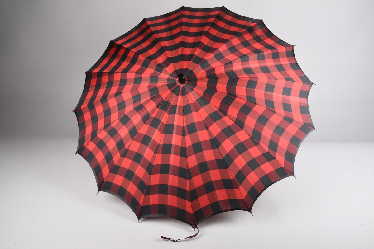 Paraply med 16 metallspilar. Stong i tre. Trekt med stoff i kunstfiber, rutemønster. Handtak i tre med hempe i tekstil til å ta inn på handa. Hempe for lukking av paraplyen.